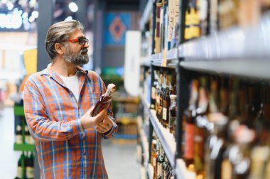 Bir adam süpermarketin rafından alkollü içecekler alır. Alkol almak için markete gidiyorum..