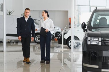 Araba galerisinde genç ve çekici bir kadın satıcı erkek müşterisine bir araba gösteriyor..