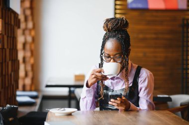 Kafedeki genç Afrikalı kadın kahve içiyor ve cep telefonu kullanıyor..