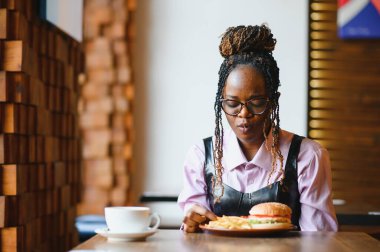 Kafede hamburger ve patates yiyen Afrikalı bir kız. Fast food konsepti.