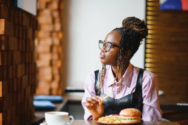 Kafede hamburger ve patates yiyen Afrikalı bir kız. Fast food konsepti.