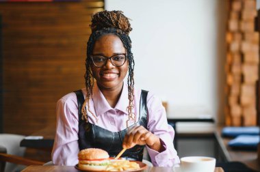 Kafede hamburger ve patates yiyen Afrikalı bir kız. Fast food konsepti.