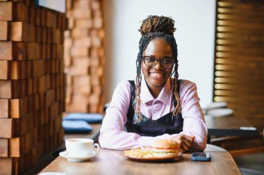 Kafede hamburger ve patates yiyen Afrikalı bir kız. Fast food konsepti.