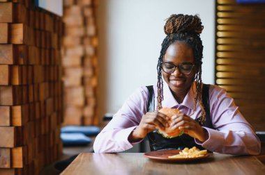 Afro saçlı Afrikalı bir kadın leziz bir klasik burgerle patates kızartması yiyor. Hile Yemeği.