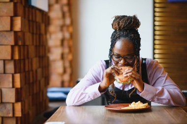Kafede hamburger ve patates yiyen Afrikalı bir kız. Fast food konsepti.