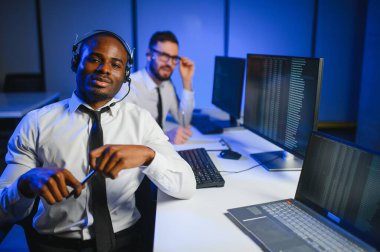 Mutlu Güvenilir Teknik Müşteri Destek Uzmanı Meslektaşlar ve Ekranlarla Dolu Gözetleme ve Kontrol Odasında Kulaklık Çağrısı Yapıyor.