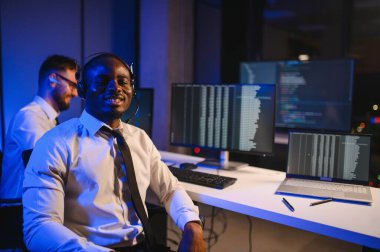 Mutlu Güvenilir Teknik Müşteri Destek Uzmanı Meslektaşlar ve Ekranlarla Dolu Gözetleme ve Kontrol Odasında Kulaklık Çağrısı Yapıyor.