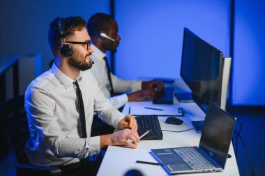 Mutlu Güvenilir Teknik Müşteri Destek Uzmanı Meslektaşlar ve Ekranlarla Dolu Gözetleme ve Kontrol Odasında Kulaklık Çağrısı Yapıyor.