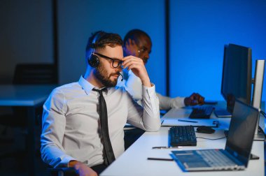 Mutlu Güvenilir Teknik Müşteri Destek Uzmanı Meslektaşlar ve Ekranlarla Dolu Gözetleme ve Kontrol Odasında Kulaklık Çağrısı Yapıyor.