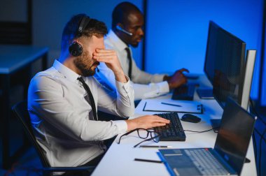 Mutlu Güvenilir Teknik Müşteri Destek Uzmanı Meslektaşlar ve Ekranlarla Dolu Gözetleme ve Kontrol Odasında Kulaklık Çağrısı Yapıyor.