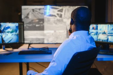 Afro-Amerikan güvenlik operatörünün arka görüntüsü. Güvenlik kamerası görüntülerini gösteren ekranları olan bir bilgisayar kullanın. .