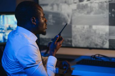 Telsizle konuşurken masaüstü bilgisayar ekranına bakan Afrikalı Amerikalı güvenlik görevlisi.