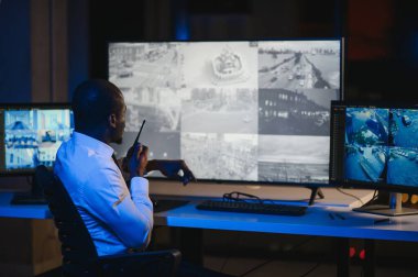 Afro-Amerikan güvenlik operatörünün arka görüntüsü. Güvenlik kamerası görüntülerini gösteren ekranları olan bir bilgisayar kullanın. .