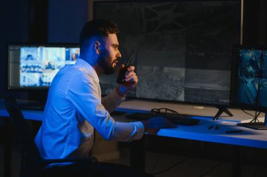 Erkek güvenlik görevlisi, bilgisayardaki birden fazla CCTV görüntüsünü izlerken telsizden konuşuyor..