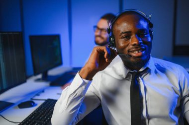 Mutlu Güvenilir Teknik Müşteri Destek Uzmanı Meslektaşlar ve Ekranlarla Dolu Gözetleme ve Kontrol Odasında Kulaklık Çağrısı Yapıyor.