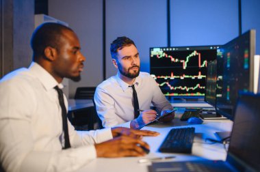 İki işadamı, finans analisti ya da yatırım danışmanı ellerinde polis bilgisayarları ve belgelerle büro masasında oturuyorlar.