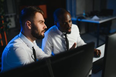 İki işadamı, finans analisti ya da yatırım danışmanı ellerinde polis bilgisayarları ve belgelerle büro masasında oturuyorlar.