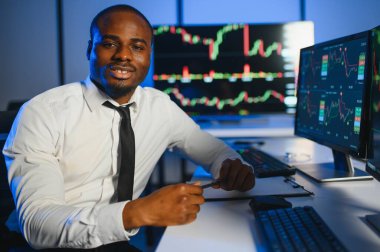 Afro-amerikan borsa bürosunda çalışan borsacı veya komisyoncu. Arka planda veri, saat numaraları ve grafikleri gösteren monitörlerin bilgisayarını kullanıyor..