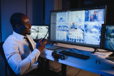 Telsizle konuşurken masaüstü bilgisayar ekranına bakan Afrikalı Amerikalı güvenlik görevlisi.