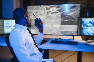 Telsizle konuşurken masaüstü bilgisayar ekranına bakan Afrikalı Amerikalı güvenlik görevlisi.