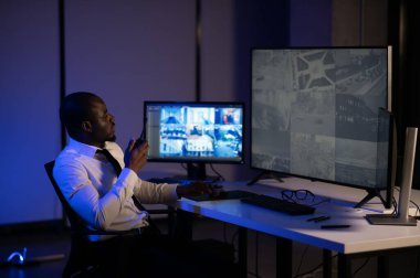 Telsizle konuşurken masaüstü bilgisayar ekranına bakan Afrikalı Amerikalı güvenlik görevlisi.