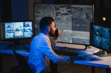 Erkek güvenlik görevlisi, bilgisayardaki birden fazla CCTV görüntüsünü izlerken telsizden konuşuyor..