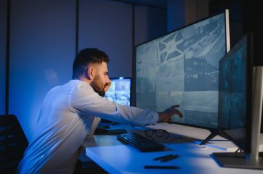 Erkek güvenlik görevlisi, bilgisayardaki birden fazla CCTV görüntüsünü izlerken telsizden konuşuyor..