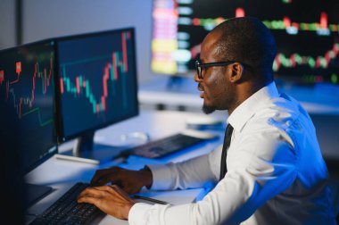 Afro-amerikan borsa bürosunda çalışan borsacı veya komisyoncu. Arka planda veri, saat numaraları ve grafikleri gösteren monitörlerin bilgisayarını kullanıyor..