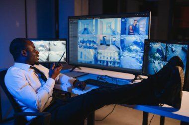 Telsizle konuşurken masaüstü bilgisayar ekranına bakan Afrikalı Amerikalı güvenlik görevlisi.