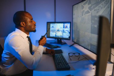 Telsizle konuşurken masaüstü bilgisayar ekranına bakan Afrikalı Amerikalı güvenlik görevlisi.