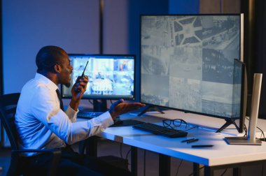 Telsizle konuşurken masaüstü bilgisayar ekranına bakan Afrikalı Amerikalı güvenlik görevlisi.