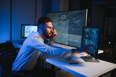 Erkek güvenlik görevlisi, bilgisayardaki birden fazla CCTV görüntüsünü izlerken telsizden konuşuyor..