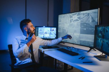 Erkek güvenlik görevlisi, bilgisayardaki birden fazla CCTV görüntüsünü izlerken telsizden konuşuyor..