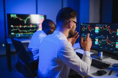 İki işadamı, finans analisti ya da yatırım danışmanı ellerinde polis bilgisayarları ve belgelerle büro masasında oturuyorlar.