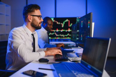 İki işadamı, finans analisti ya da yatırım danışmanı ellerinde polis bilgisayarları ve belgelerle büro masasında oturuyorlar.