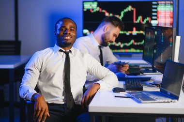 İki işadamı, finans analisti ya da yatırım danışmanı ellerinde polis bilgisayarları ve belgelerle büro masasında oturuyorlar.