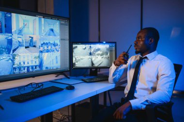 Ciddi bir Afrikalı Amerikalı güvenlik görevlisi güvenlik odasında güvenlik kamerası görüntülerini izlerken bilgisayar ekranından otel hayatını izliyor.