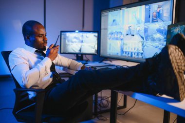 Ciddi bir Afrikalı Amerikalı güvenlik görevlisi güvenlik odasında güvenlik kamerası görüntülerini izlerken bilgisayar ekranından otel hayatını izliyor.