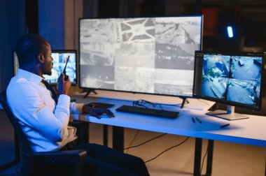 Telsizle konuşurken masaüstü bilgisayar ekranına bakan Afrikalı Amerikalı güvenlik görevlisi.