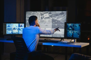 Erkek güvenlik görevlisi, bilgisayardaki birden fazla CCTV görüntüsünü izlerken telsizden konuşuyor..