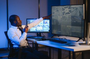 Telsizle konuşurken masaüstü bilgisayar ekranına bakan Afrikalı Amerikalı güvenlik görevlisi.