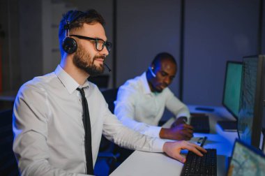 Mutlu Güvenilir Teknik Müşteri Destek Uzmanı Meslektaşlar ve Ekranlarla Dolu Gözetleme ve Kontrol Odasında Kulaklık Çağrısı Yapıyor.