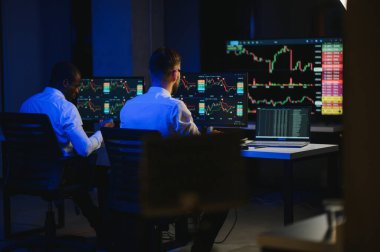 İki işadamı, finans analisti ya da yatırım danışmanı ellerinde polis bilgisayarları ve belgelerle büro masasında oturuyorlar.