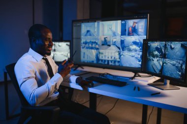 Ciddi bir Afrikalı Amerikalı güvenlik görevlisi güvenlik odasında güvenlik kamerası görüntülerini izlerken bilgisayar ekranından otel hayatını izliyor.