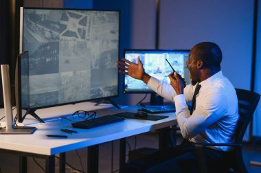 Ciddi bir Afrikalı Amerikalı güvenlik görevlisi güvenlik odasında güvenlik kamerası görüntülerini izlerken bilgisayar ekranından otel hayatını izliyor.