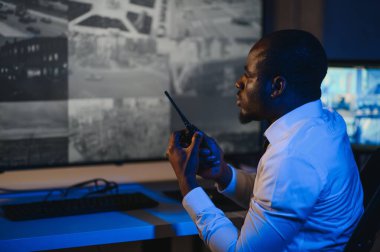 Telsizle konuşurken masaüstü bilgisayar ekranına bakan Afrikalı Amerikalı güvenlik görevlisi.