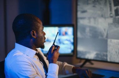Telsizle konuşurken masaüstü bilgisayar ekranına bakan Afrikalı Amerikalı güvenlik görevlisi.