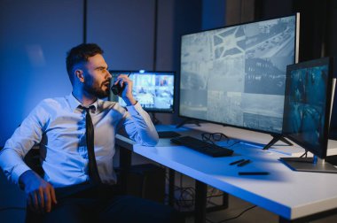 Erkek güvenlik görevlisi, bilgisayardaki birden fazla CCTV görüntüsünü izlerken telsizden konuşuyor..