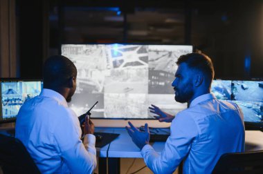 İki güvenlik görevlisi iş yerinde bilgisayar monitörlerinin önünde oturuyorlar ve monitörlerdeki güvenlik kameralarını izliyorlar.