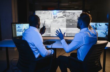 İki güvenlik görevlisi iş yerinde bilgisayar monitörlerinin önünde oturuyorlar ve monitörlerdeki güvenlik kameralarını izliyorlar.